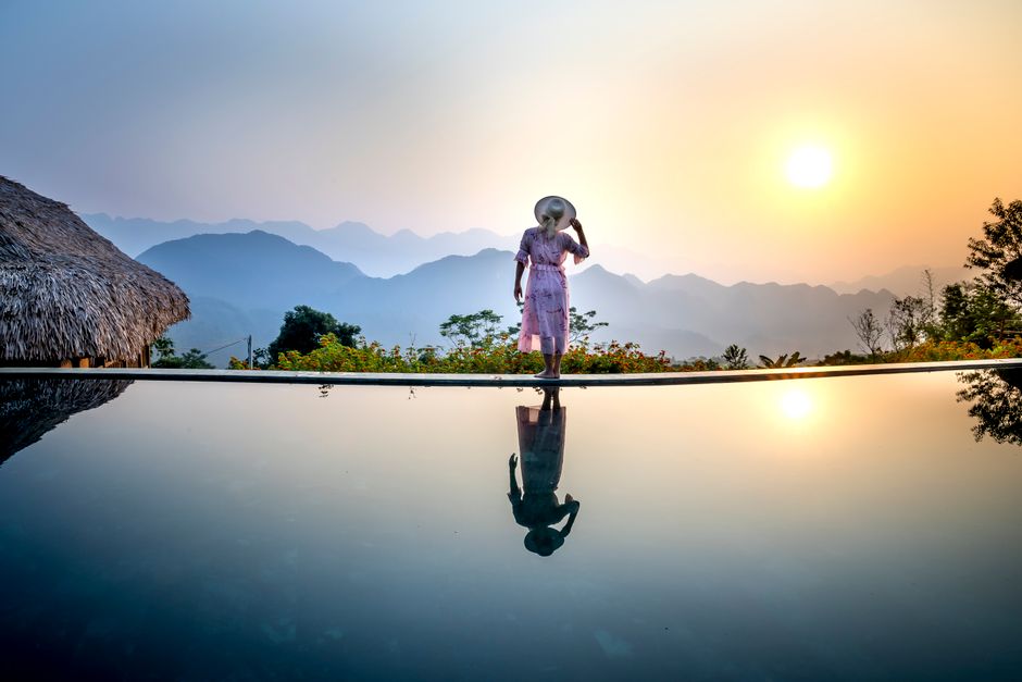 Back view of full body anonymous female in romantic dress and straw hat standing on poolside and looking away while enjoying sundown in amazing terrain